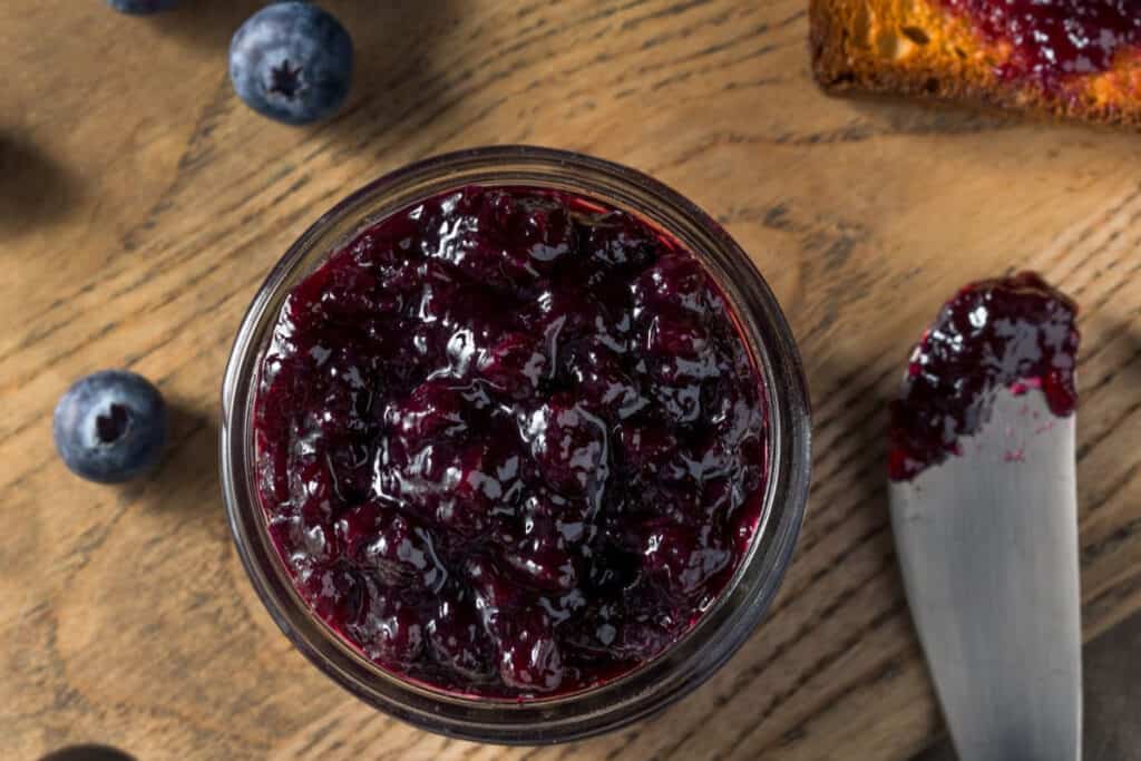An open jar of blueberry jam seen from above with a spreader on a board with fresh blueberries.