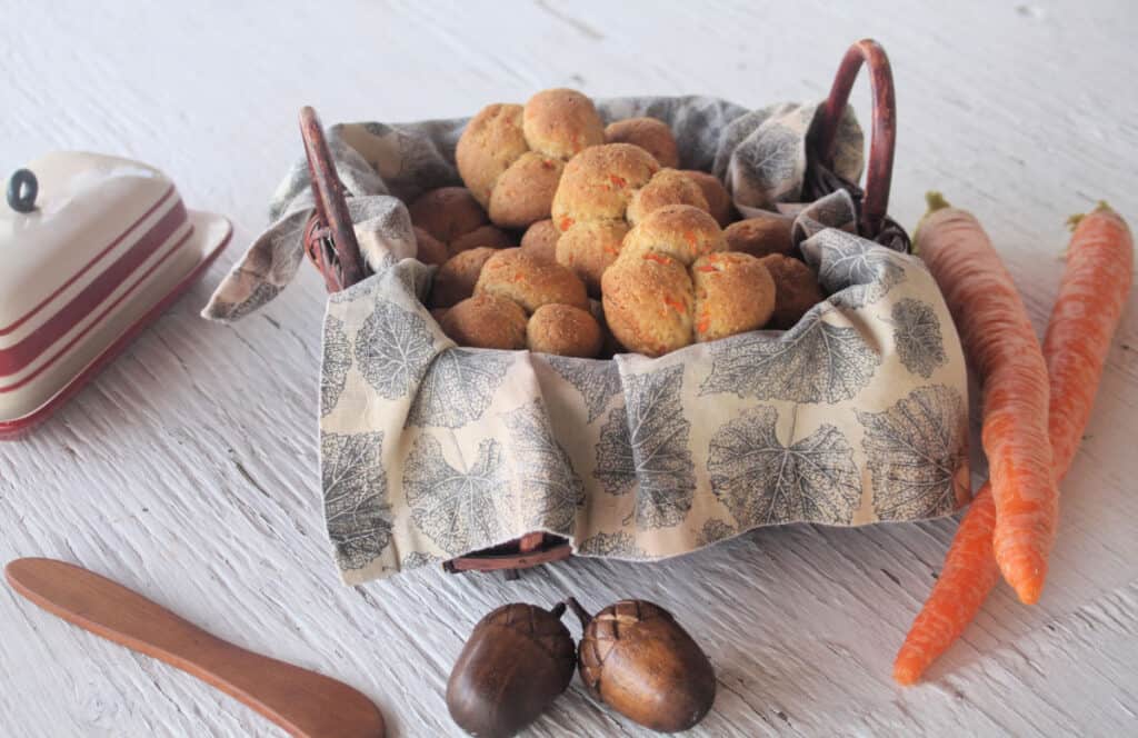 A basket of carrot cloverleaf rolls surrounded by a butter dish and fresh carrots. 