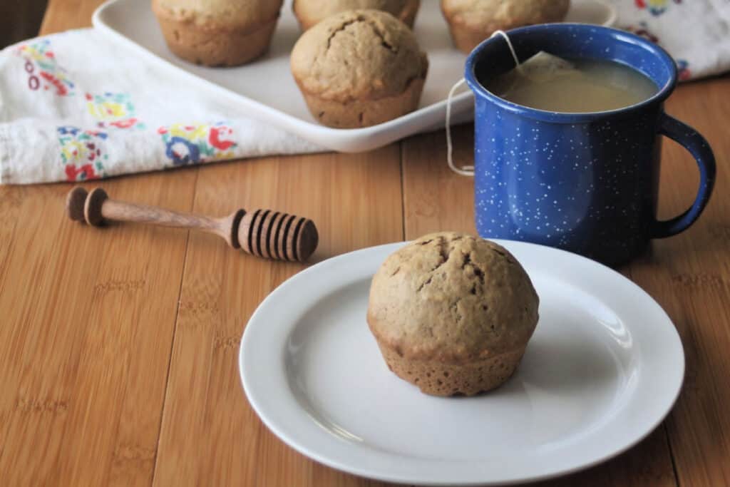 A chai muffin on a white plate surrounded my more muffins, a honey dipper, and a cup of tea.