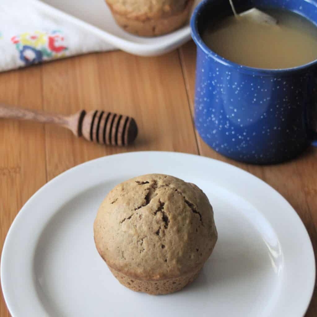 Chai muffin on a white plate surrounded by a blue cup of tea and honey dipper.