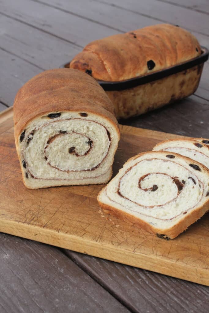 A loaf and slices of cinnamon raisin swirl bread on a wooden cutting board.