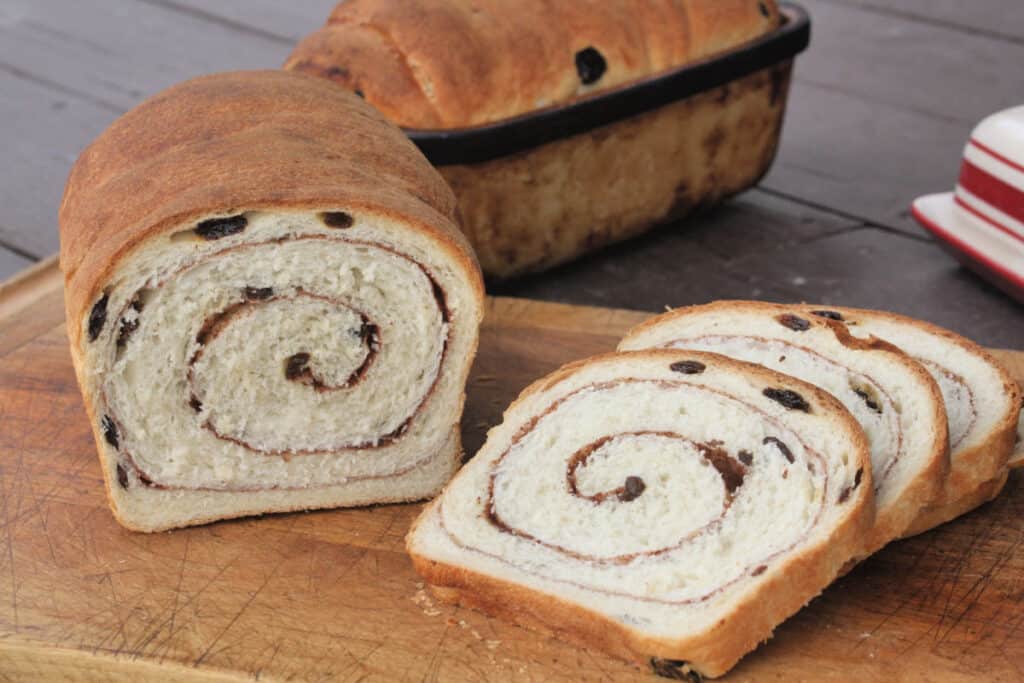 A loaf and slices of cinnamon raisin swirl bread on a wooden cutting board.