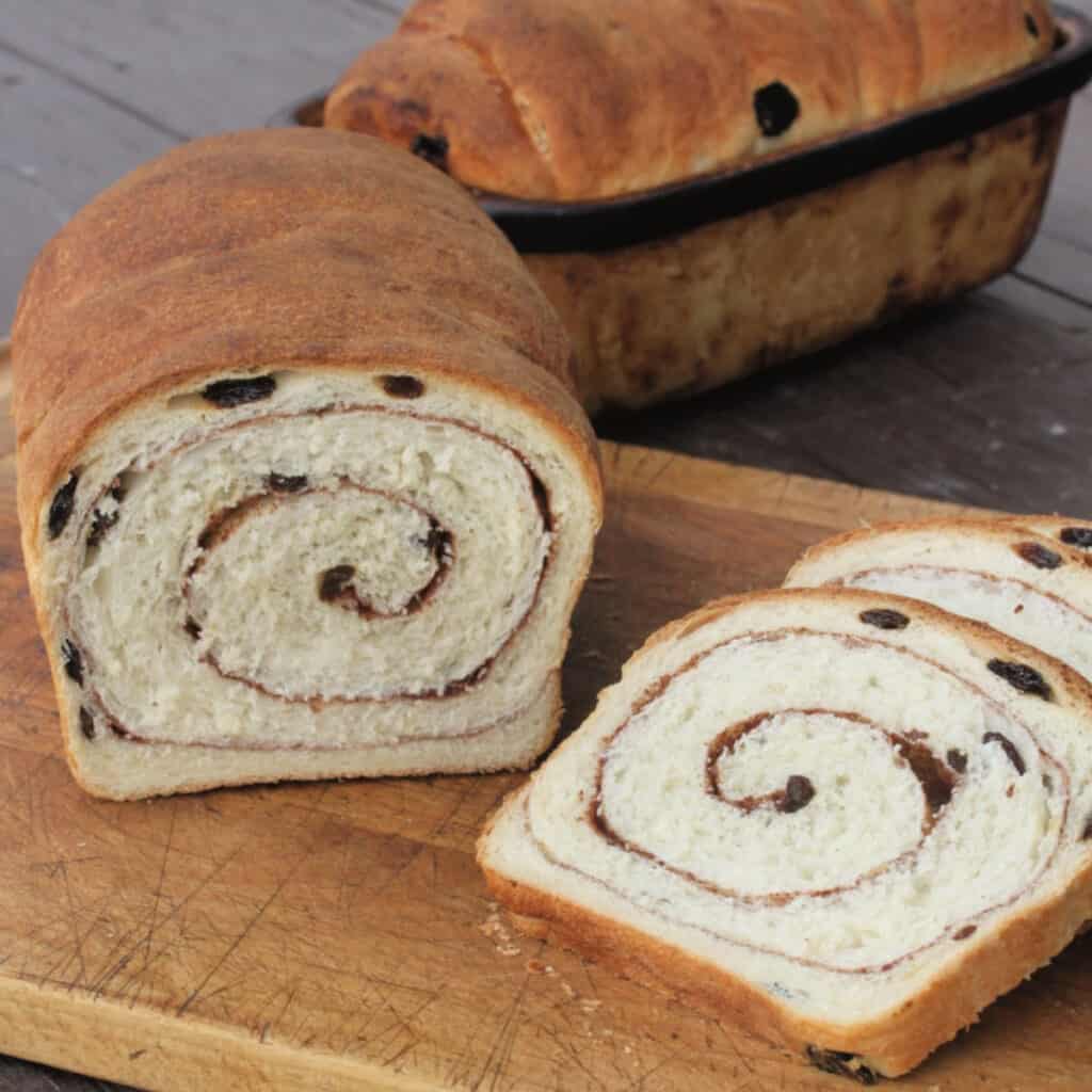 A loaf and slices of cinnamon raisin swirl bread on a wooden cutting board.