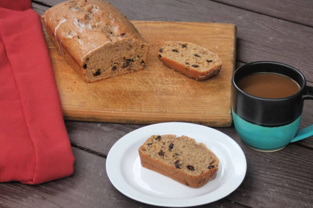 A slice of cinnamon raisin quick bread on a white plate with a cup of coffee and entire loaf of bread on a wooden cutting board.