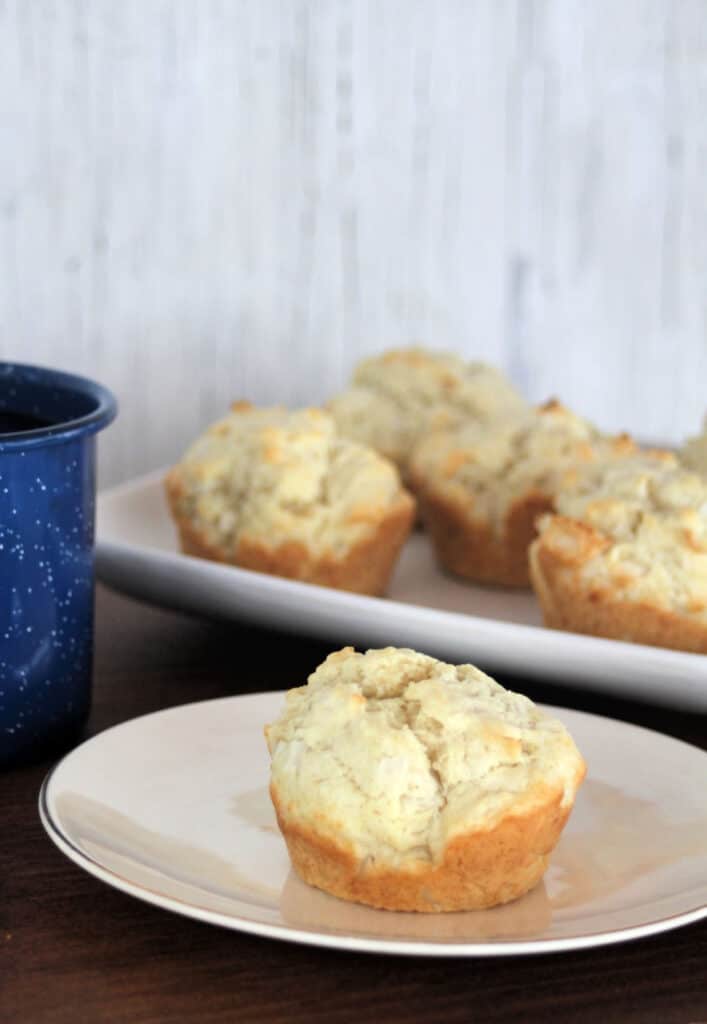 A coconut muffin on a white plate sitting next to a blue coffee cup and in front of a platter of muffins.