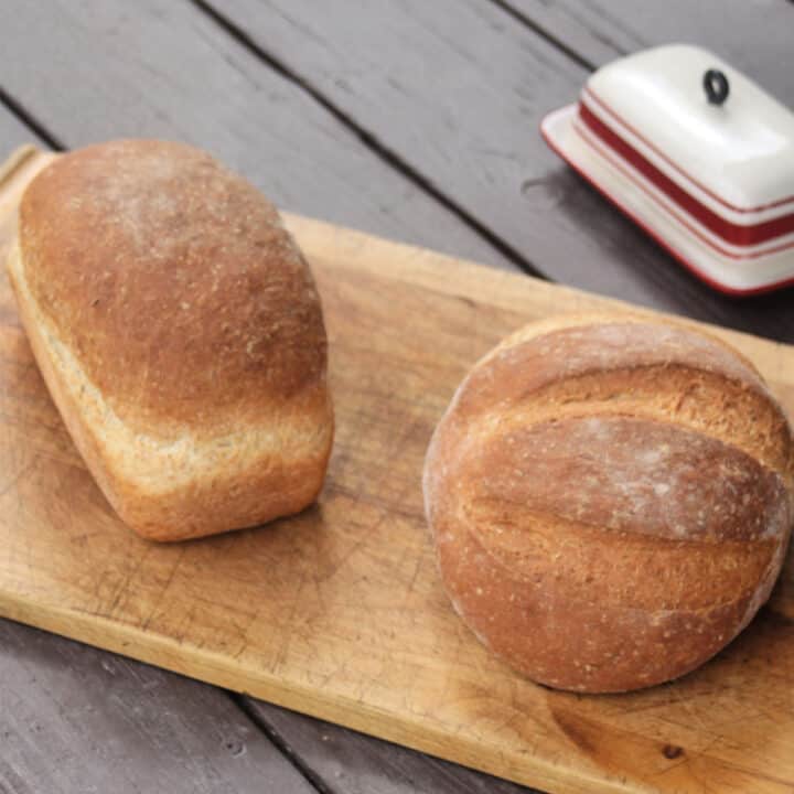 2 loaves of honey wheat bran bread on a wooden cutting board with a butter dish behind it.