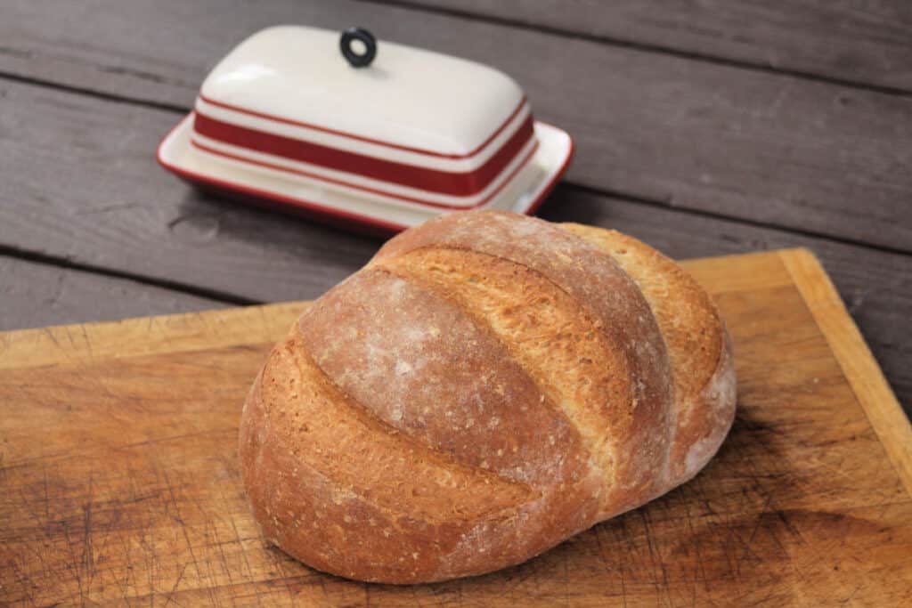 A loaf of honey wheat bran bread on a cutting board with butter dish sitting behind it.