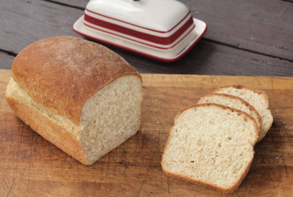 A loaf of honey wheat bran bread on a board sitting next to slices with a butter dish behind it. 