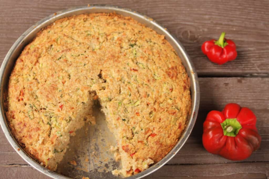 A pan of zucchini cornbread with a slice missing and 2 red peppers sitting to the right.