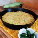 A cast iron skillet full of cornbread sits on a yellow cloth. A bowl of hot peppers sits in front of it, a whole fresh zucchini behind it. Text overlay reads: Zucchini Cornbread.