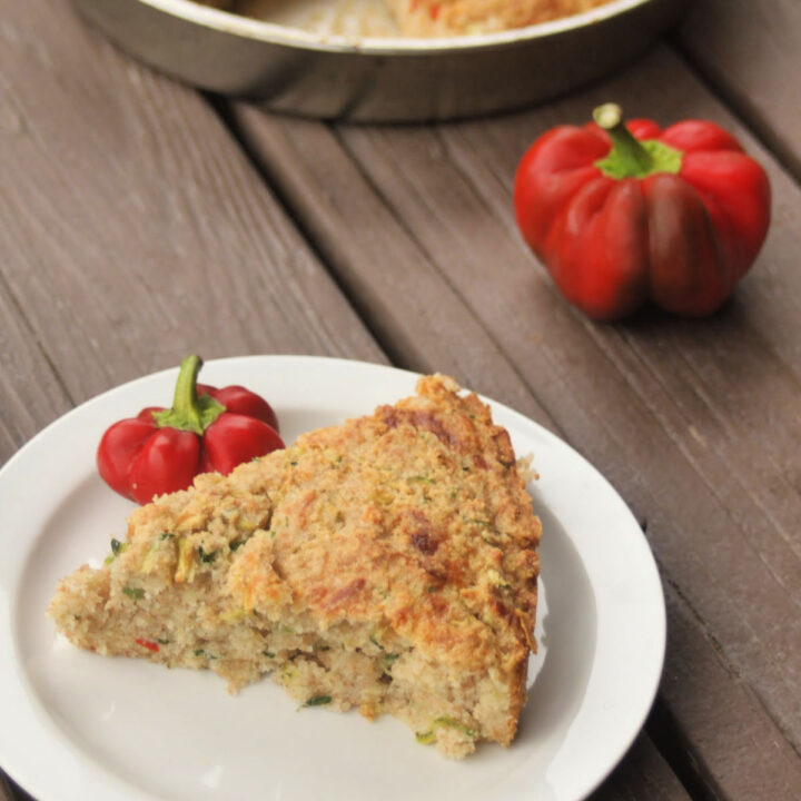 A slice of zucchini cornbread on a white plate surrounded by fresh hot red peppers.