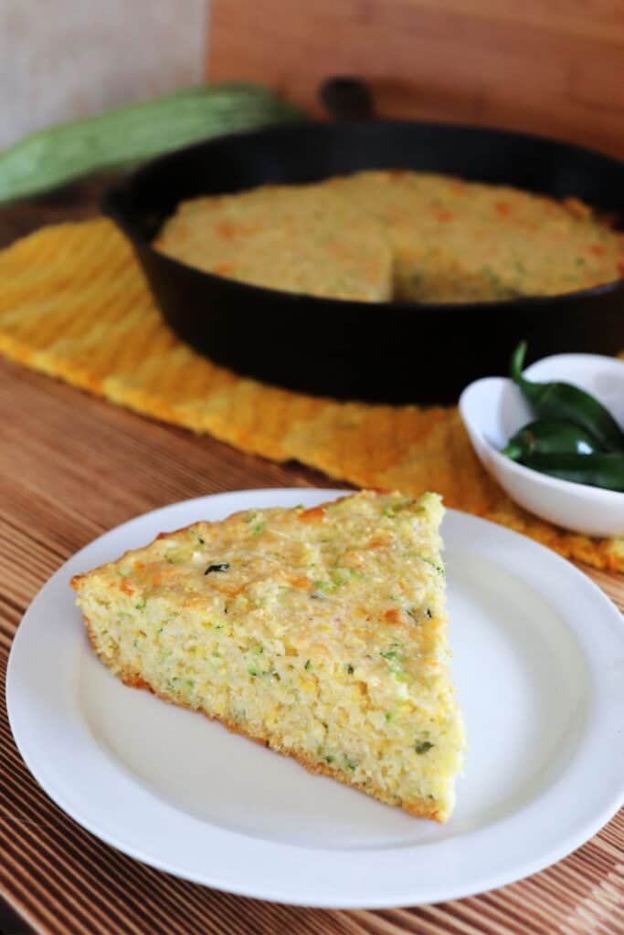 A slice of zucchini cornbread on a plate with a bowl of hot peppers and cast iron skillet with more cornbread in the background.