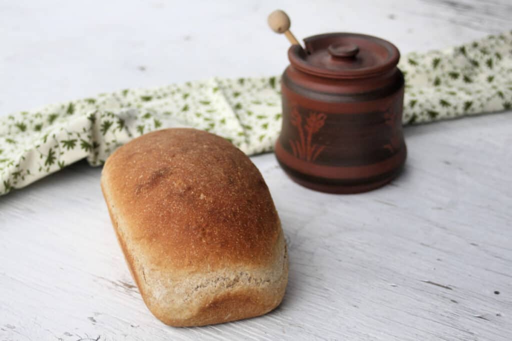 A loaf of honey graham bread on a white board with a clay pot and green and white towel.