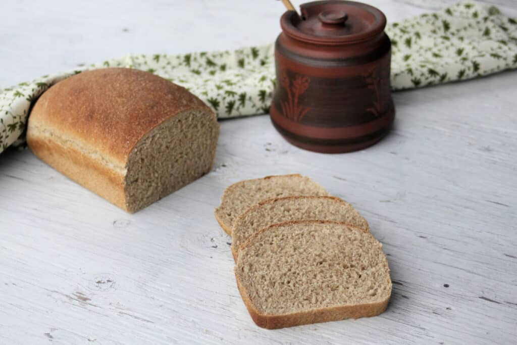 Slices of honey graham bread sitting next to a loaf with a jar of honey and green and white towel.