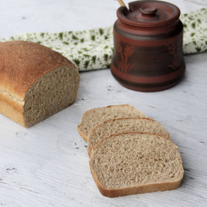 Slices of honey graham bread sitting next to a loaf with a jar of honey and green and white towel.