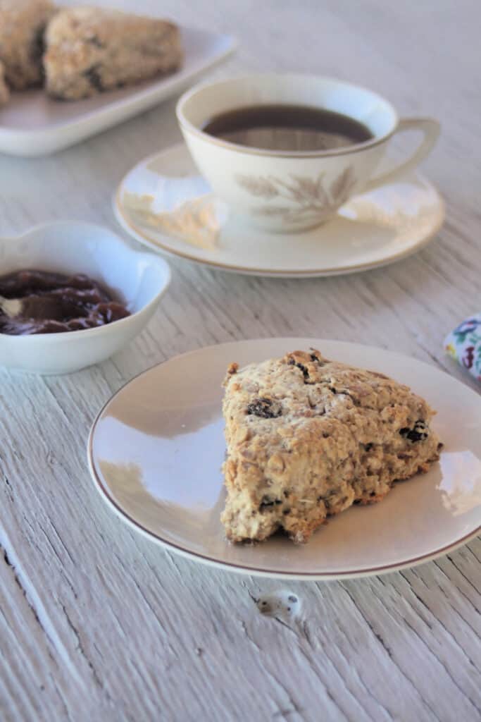 A maple muesli scone on a white plate with a cup of tea, a bowl of jam, and a cloth napkin. 