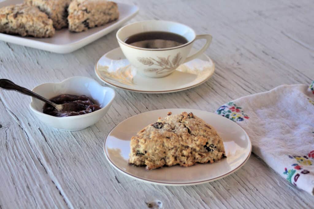 A maple muesli scone on a white plate with a cup of tea, a bowl of jam, and a cloth napkin. 