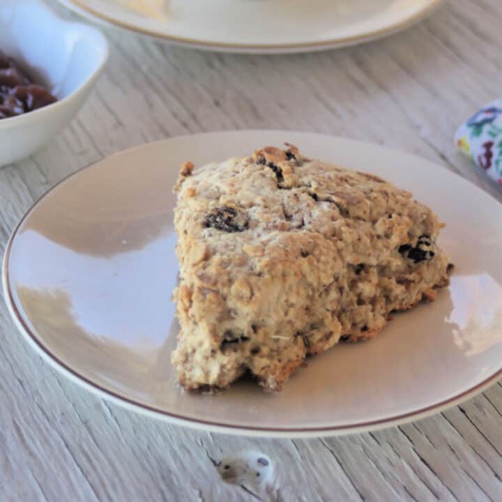 A maple muesli scone on a white plate in front of a bowl of jam with a napkin.