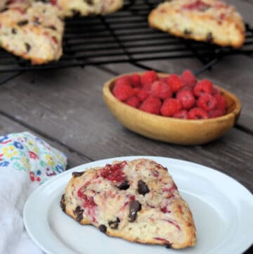 A raspberry scone on a white plate sitting next to a napkin. In the background is a bowl of fresh raspberries and more scones on a wire rack.