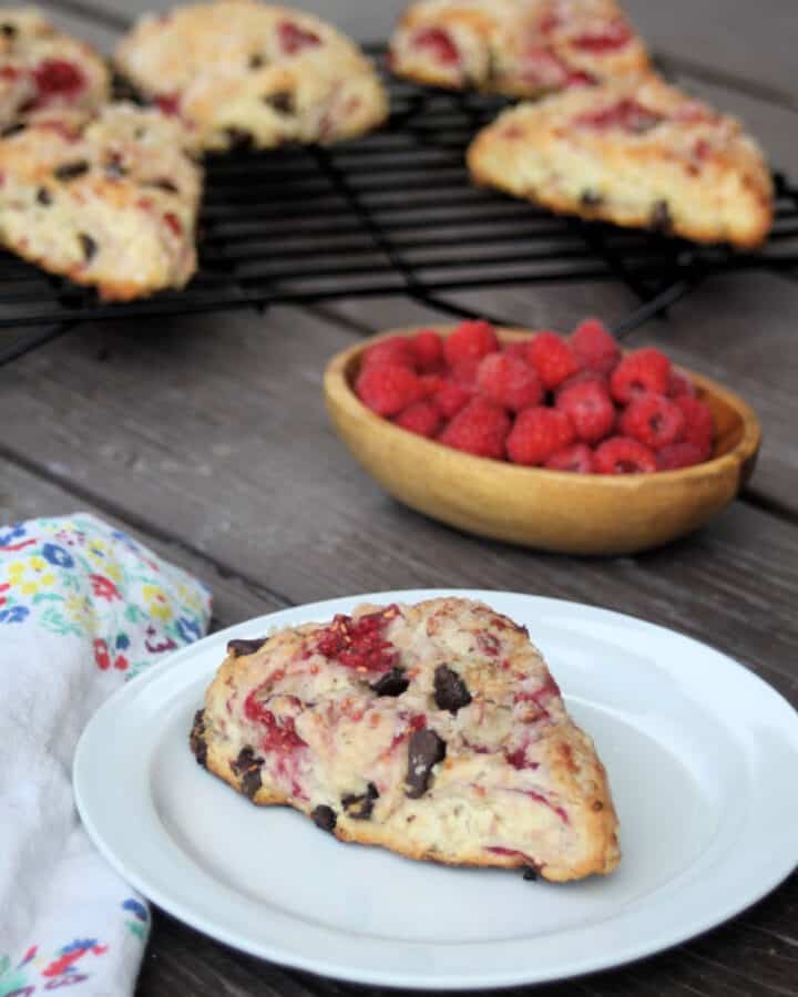 A raspberry scone on a white plate sitting next to a napkin. In the background is a bowl of fresh raspberries and more scones on a wire rack.