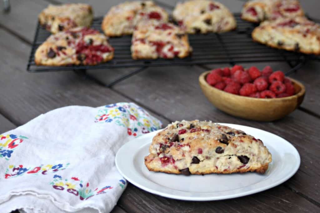 A raspberry scone on a white plate sitting next to a napkin. In the background is a bowl of fresh raspberries and more scones on a wire rack.