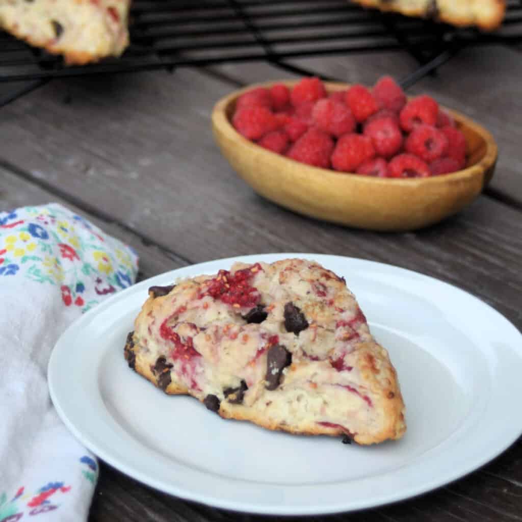 A raspberry chocolate chip scone on a white plate with a napkin and bowl of fresh rapsberries.