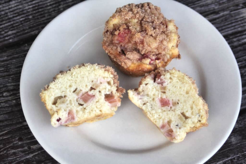 A sliced rhubarb muffin on a white plate sitting in front of a whole muffin.