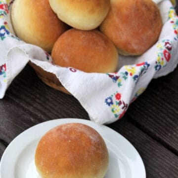 A yogurt dinner roll sitting on a white plate in front of a basket full of more rolls.