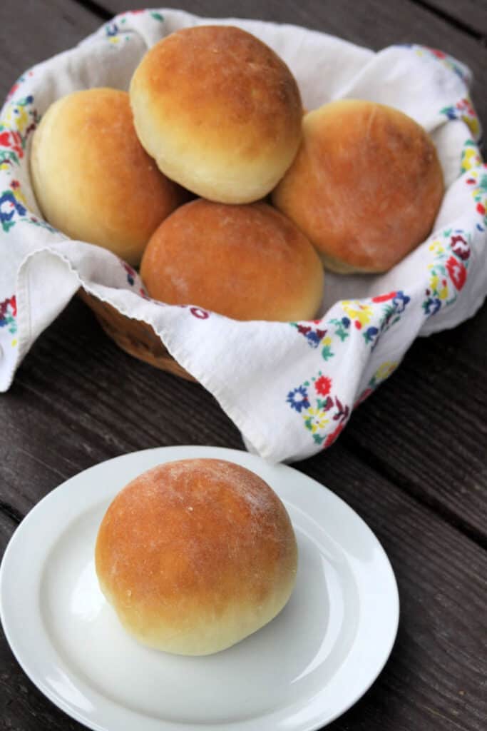 A yogurt dinner roll sitting on a white plate in front of a basket full of more rolls.