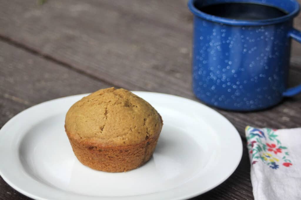 A coffee muffin on a white plate with a floral napkin and blue tin cup full of coffee next to it on the table.