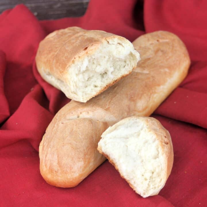 A loaf of french bread sitting on a red cloth with another torn loaf of bread sitting on top.