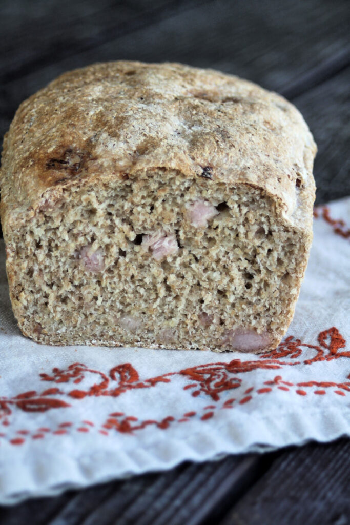 A loaf of ham bread with the end cut off exposing bits of ham inside the bread sitting on an embroidered napkin. 