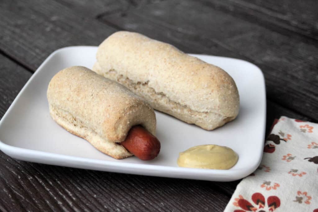 Two homemade pigs in a blanket on a white plate with mustard and napkin sitting beside the plate.