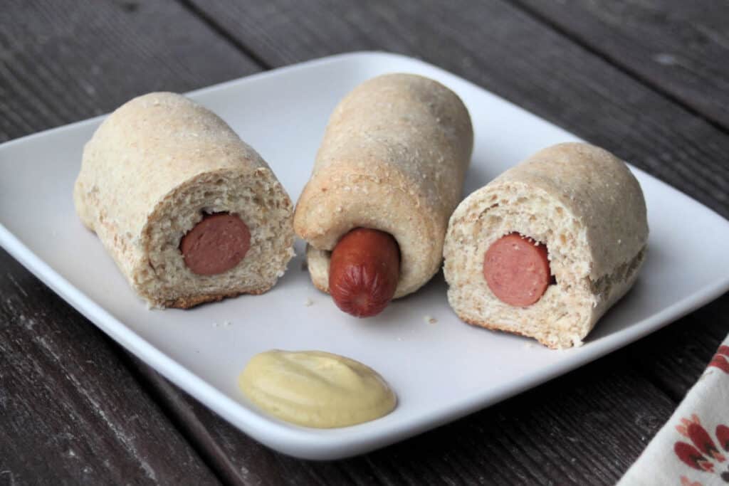 Homemade pigs in a blanket - 1 whole and 1 sliced in half - on a white plate with mustard.