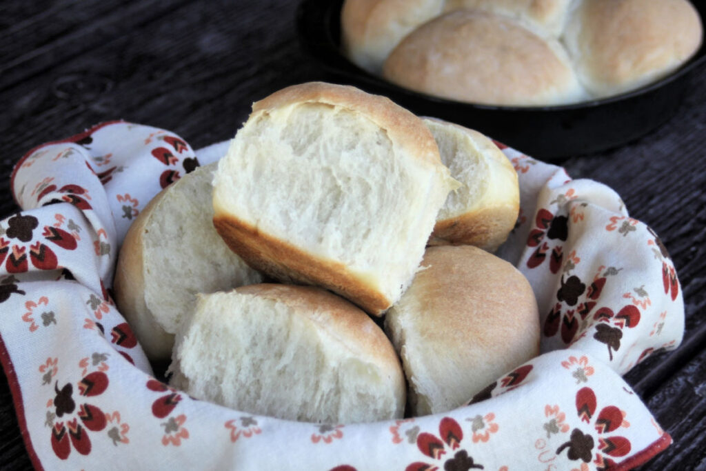 A napkin lined basket full of homemade yeast rolls with a close up of the one on top sitting in front of a metal pan full of more rolls. 