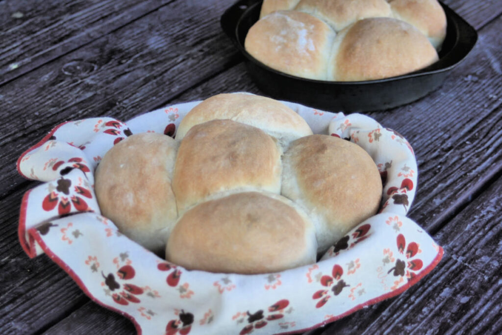 A basket of homemade yeast rolls sitting on a table in front of rolls in a metal cake pan.