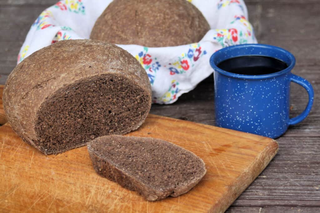 A slice of honey wheat brown bread on a wooden cutting board next to the loaf sitting with a cup of coffee on a table and another loaf sitting in a floral napkin lined basket.