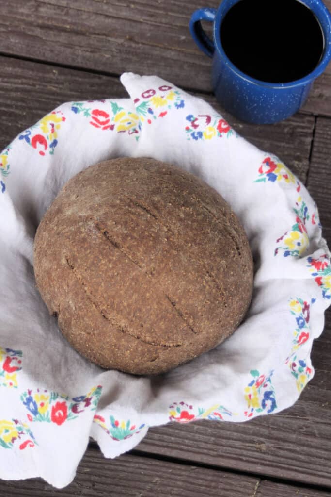 A round loaf of honey wheat brown bread in a towel lined basket with a tin cup of coffee as seen from above.