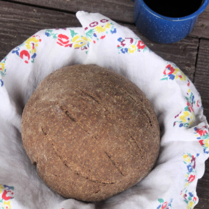 A round loaf of honey wheat brown bread in a towel lined basket as seen from above.