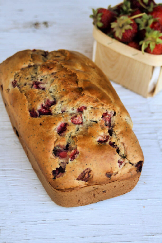 A loaf of strawberry quick bread on a white table with a basket of fresh berries behind it. 