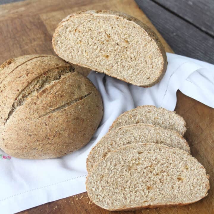 Slices of whole grain cheddar bread on a cutting board with two loaves sitting on a white cloth.