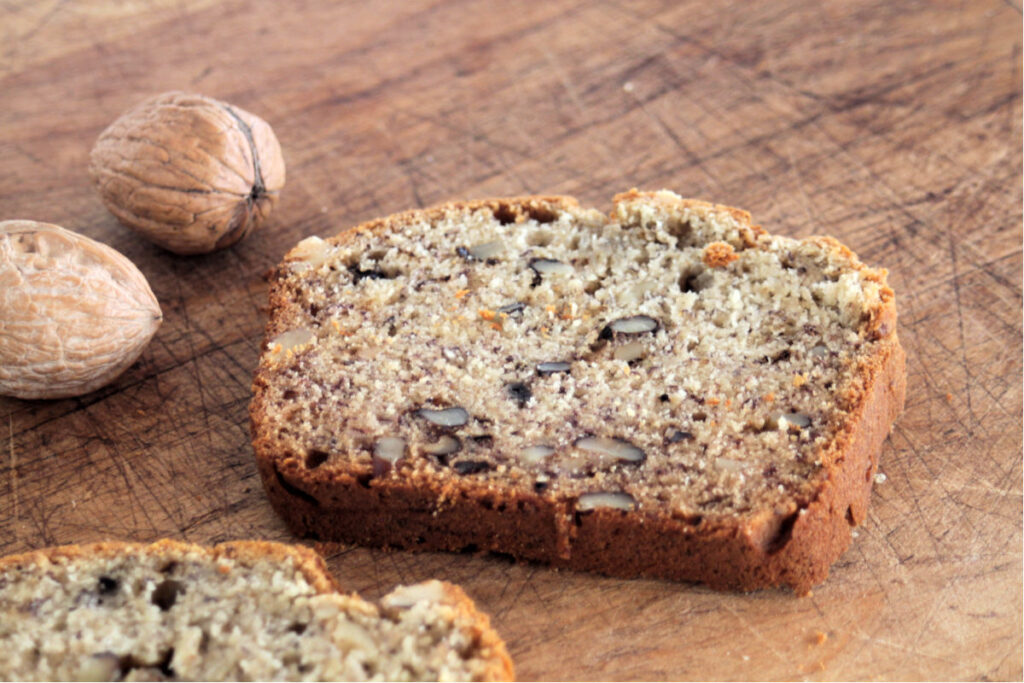 A slice of buttermilk banana bread on a cutting board with whole walnuts in their shell. 