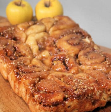 Caramel apple cinnamon rolls sitting on a wooden board with fresh apples behind it.