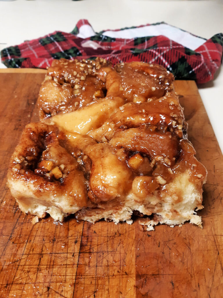 Caramel apple cinnamon rolls sitting on a wooden cutting board with a red towel behind it. 