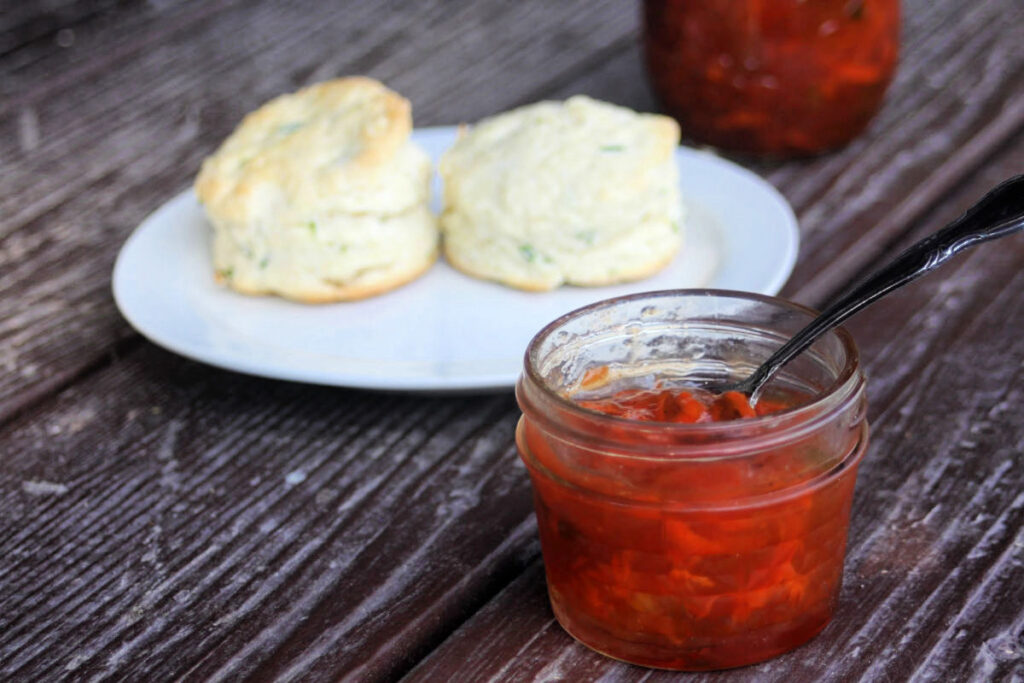 Carrot jam in an open a jar with a spoon inside it sitting in front of a white plate with 2 biscuits sitting on it.