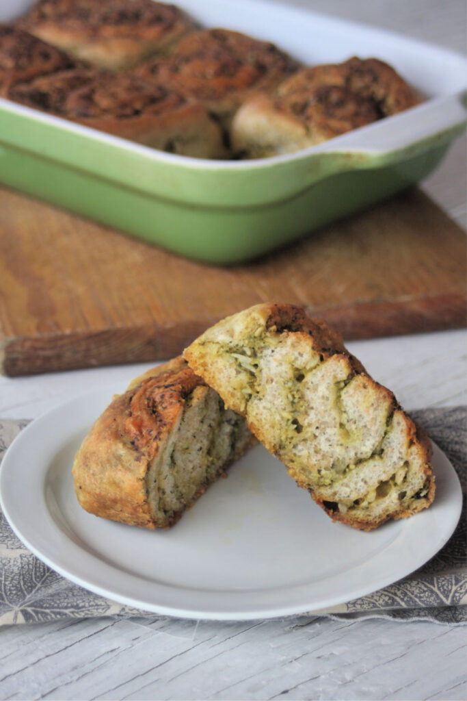 A cheesy pesto roll sliced in half on a white plate sitting on top a napkin in front of an entire pan of rolls on a wood cutting board.