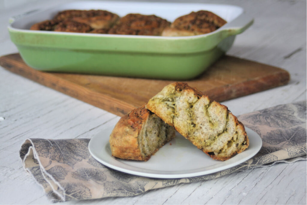 A cheesy pesto roll sliced in half on a white plate sitting on top a napkin in front of an entire pan of rolls on a wood cutting board.