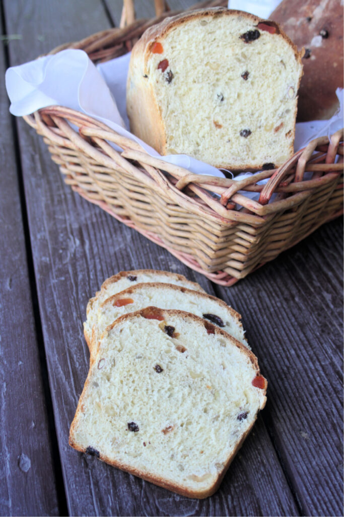 slices of harvest bread on a dark board sitting in front of a basket containing the rest of the loaf and another whole loaf of bread.