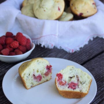A raspberry cream cheese muffin sliced in half on a white plate sitting in front of a basket full of muffins and white bowl full of fresh raspberries.