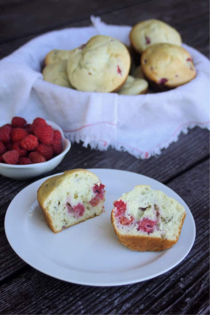 A raspberry cream cheese muffin sliced in half on a white plate sitting in front of a basket full of muffins and white bowl full of fresh raspberries.